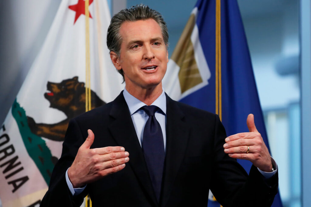 California Governor Gavin Newsom speaking at a press conference, with California state flags in the background