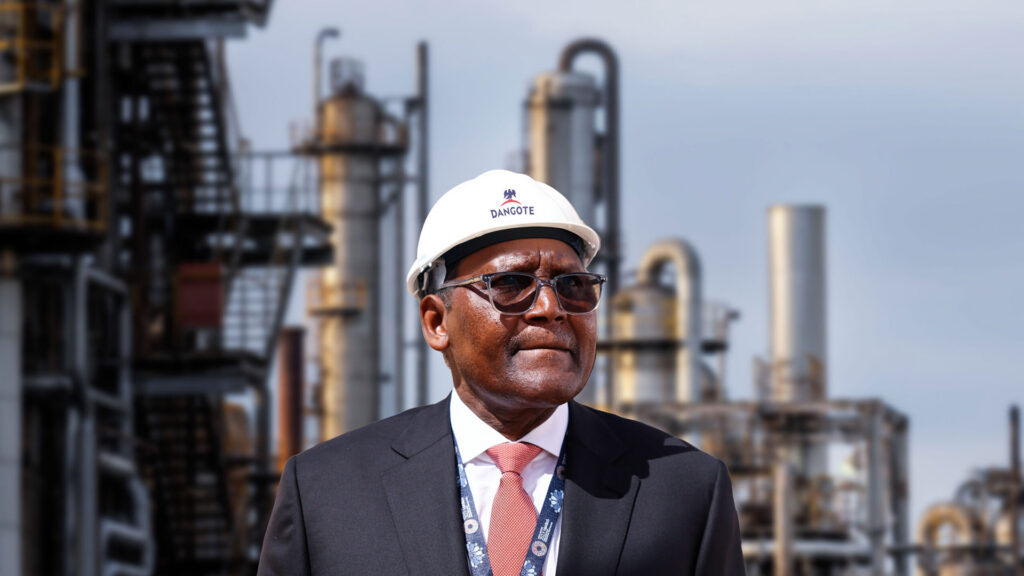 Aliko dangote wearing a hard hat and suit standing in front of industrial structures at the dangote refinery