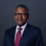 Aliko Dangote, wearing glasses and a suit with a red tie, smiling confidently in a formal portrait.