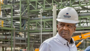 Aliko Dangote wearing a white hard hat labeled 'Dangote Fertilizer,' standing in front of industrial structures at the Dangote Fertilizer Plant.