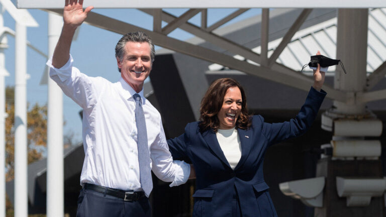 California Governor Gavin Newsom and Vice President Kamala Harris waving to a crowd during a public event, both smiling and looking energized.