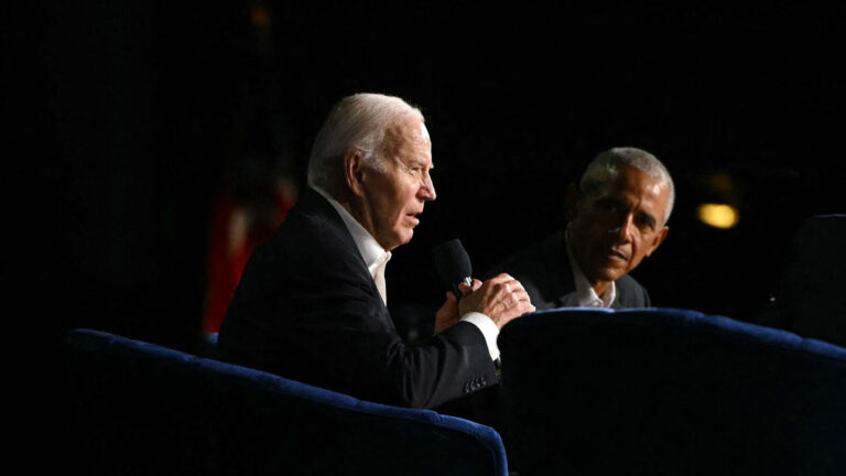 President Joe Biden speaking into a microphone with former President Barack Obama listening beside him during an event.