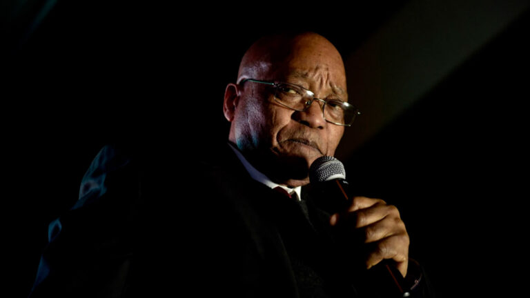 Jacob Zuma speaking into a microphone during a late-night address, wearing glasses and a dark suit.