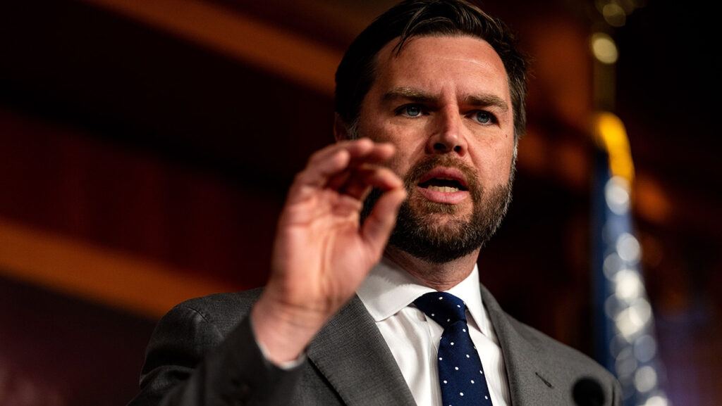 A bearded man in a suit and tie gesturing with his hand while speaking at a podium, with a partial view of an American flag in the background.