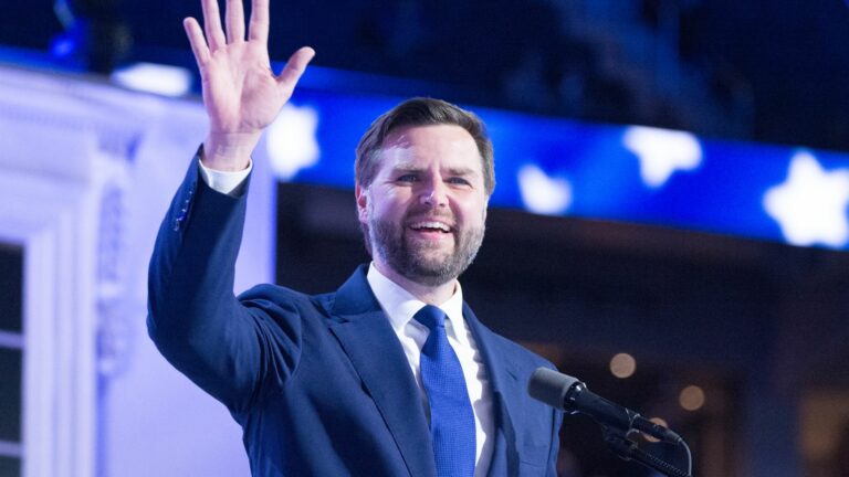 J.D. Vance, wearing a blue suit and tie, waves to the audience while speaking at the 2024 Republican National Convention. The background features a patriotic display with stars, emphasizing the event's significance.