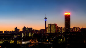 A stunning view of the Johannesburg skyline at dusk, featuring the iconic Hillbrow Tower and illuminated buildings against a vibrant sunset backdrop.