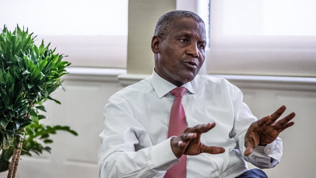 Aliko dangote wearing a white shirt and pink tie speaking and gesturing with his hands during an interview with plants in the background