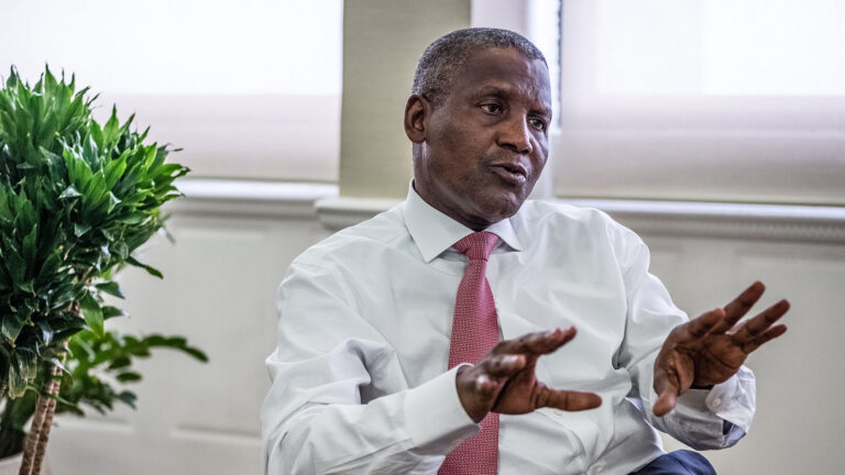 Aliko Dangote, wearing a white shirt and pink tie, speaking and gesturing with his hands during an interview, with plants in the background.
