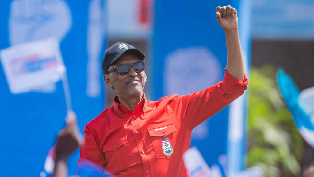 Rwandan president paul kagame wearing a red shirt and a black cap raises his fist in the air at a party rally with supporters waving flags in the background