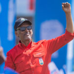 Rwandan president paul kagame wearing a red shirt and a black cap raises his fist in the air at a party rally with supporters waving flags in the background