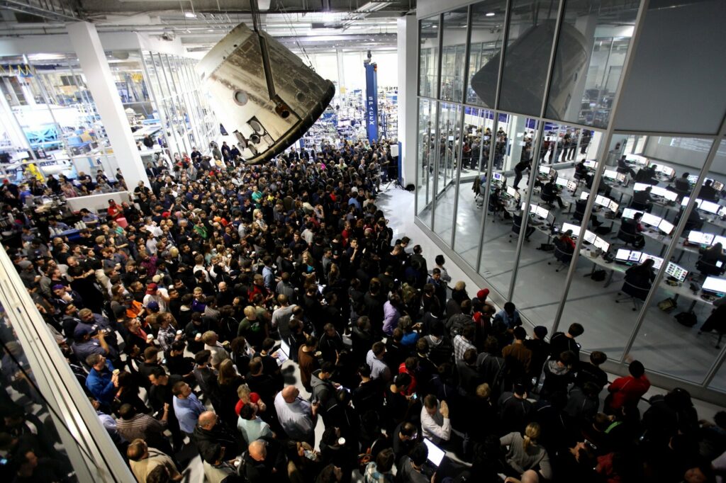 A large crowd of SpaceX employees inside the Hawthorne, California headquarters, with a spacecraft module hanging from the ceiling and glass-enclosed control rooms visible in the background.