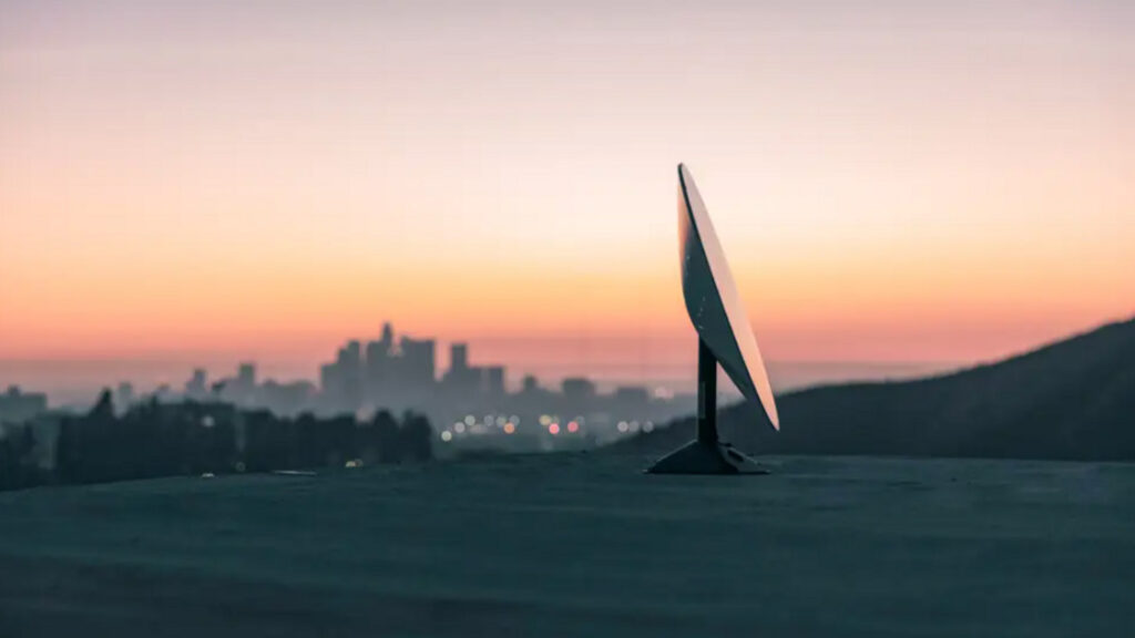 A Starlink satellite dish sits on a hilltop overlooking a city skyline at sunset, with the sky painted in warm orange and pink hues.