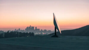 A starlink satellite dish sits on a hilltop overlooking a city skyline at sunset with the sky painted in warm orange and pink hues