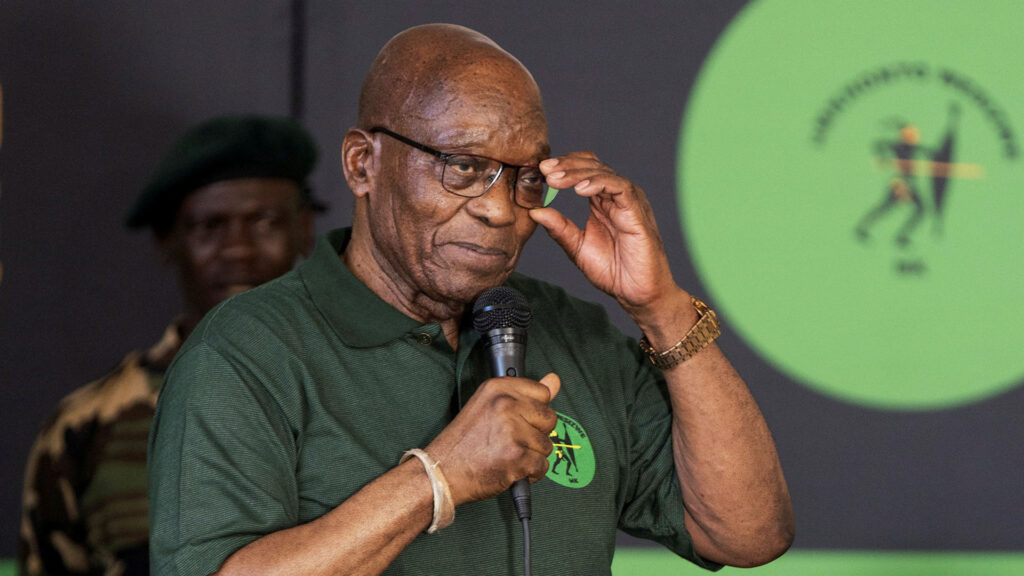 Former South African President Jacob Zuma, wearing glasses and an MK party polo shirt, speaks into a microphone at an event.