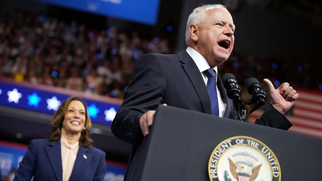 Gov. Tim Walz speaking at a podium with Vice President Kamala Harris smiling behind him at a rally.