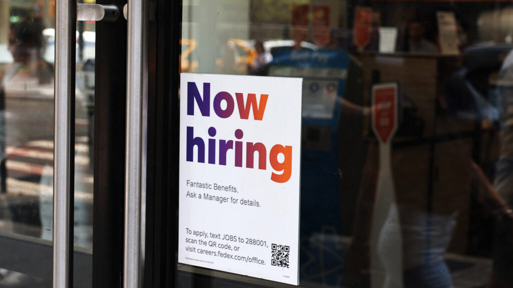 A now hiring sign posted on the glass door of a store offering details on job benefits and application instructions