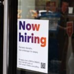 A 'Now Hiring' sign posted on the glass door of a store, offering details on job benefits and application instructions.