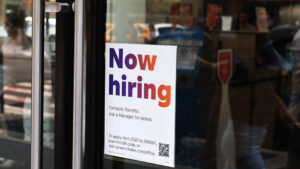 A 'Now Hiring' sign posted on the glass door of a store, offering details on job benefits and application instructions.