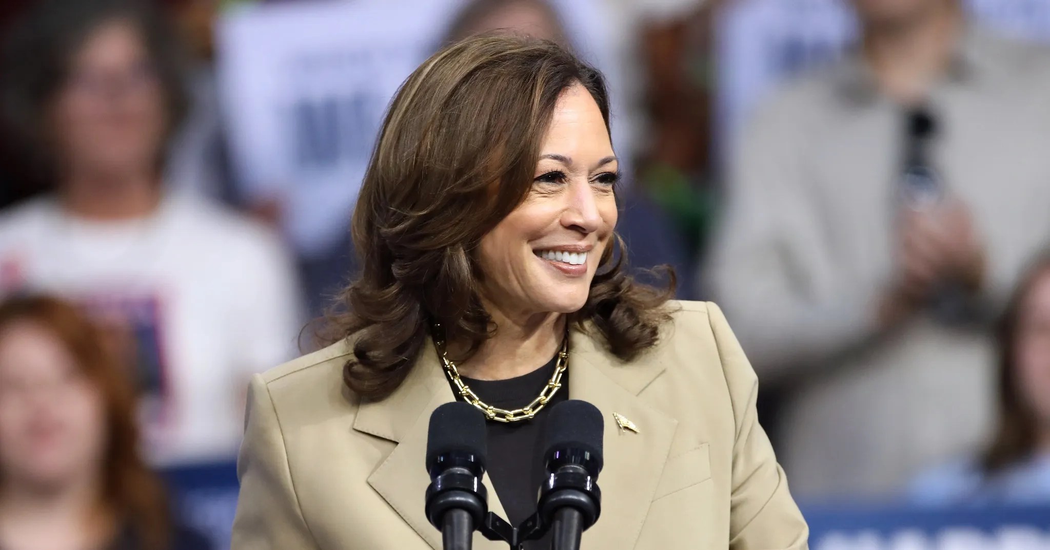 Kamala harris smiling while speaking at a podium during a political rally wearing a tan blazer and gold chain necklace with blurred supporters in the background