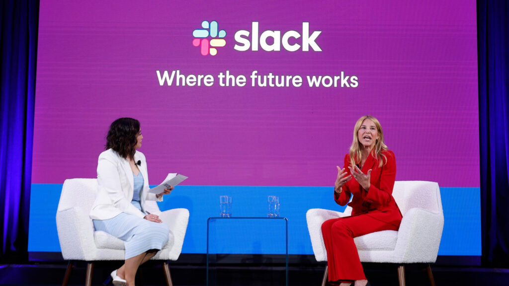 Slack ceo denise dresser speaking on stage with a moderator during a keynote event about the future of work with a large slack logo and tagline in the background