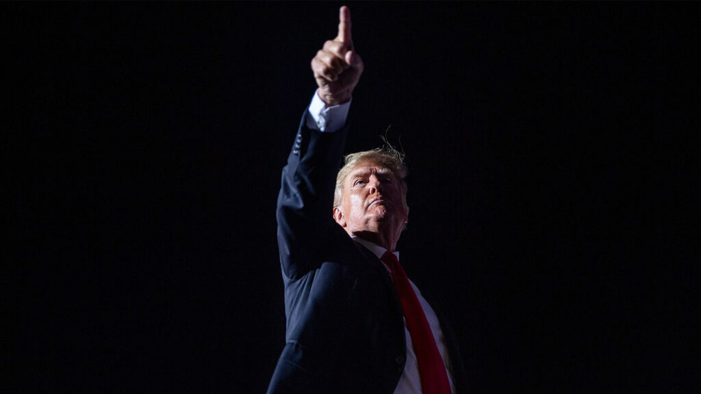 This image captures former u S President donald trump standing against a stark dark background with his hand raised in a strong pointed gesture Dressed in his usual dark suit white shirt and red tie trumps expression is determined reflecting his assertive and defiant style that has defined his public persona The dramatic lighting focuses on his face and hand casting him as a larger than life figure amid the shadows symbolizing his continued influence and uncompromising stance in american politics