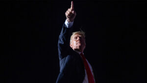 This image captures former u S President donald trump standing against a stark dark background with his hand raised in a strong pointed gesture Dressed in his usual dark suit white shirt and red tie trumps expression is determined reflecting his assertive and defiant style that has defined his public persona The dramatic lighting focuses on his face and hand casting him as a larger than life figure amid the shadows symbolizing his continued influence and uncompromising stance in american politics