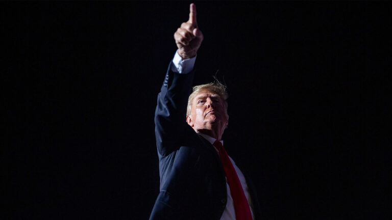 This image captures former U.S. President Donald Trump standing against a stark, dark background, with his hand raised in a strong, pointed gesture. Dressed in his usual dark suit, white shirt, and red tie, Trump’s expression is determined, reflecting his assertive and defiant style that has defined his public persona. The dramatic lighting focuses on his face and hand, casting him as a larger-than-life figure amid the shadows, symbolizing his continued influence and uncompromising stance in American politics.