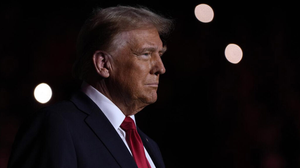 Donald trump in a dark suit and red tie shown in profile against a black background with soft distant lights