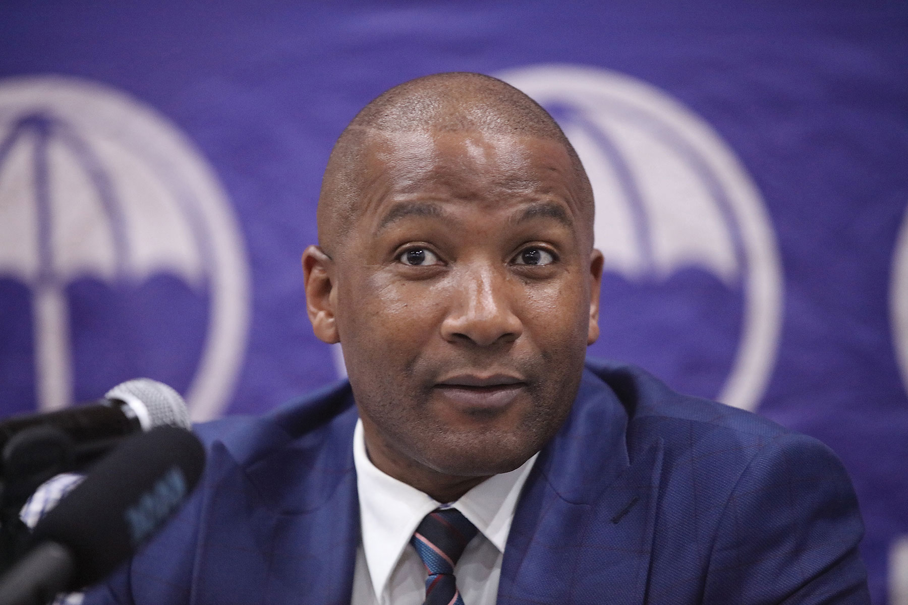 President duma boko at a press conference wearing a blue suit and tie with microphones in the foreground and a purple backdrop featuring a party logo