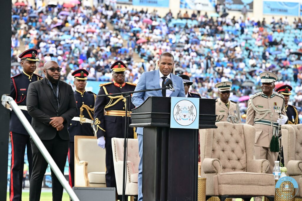 President duma boko at a podium delivering his inaugural speech with botswanas security officials and dignitaries standing beside him and a large crowd in the background