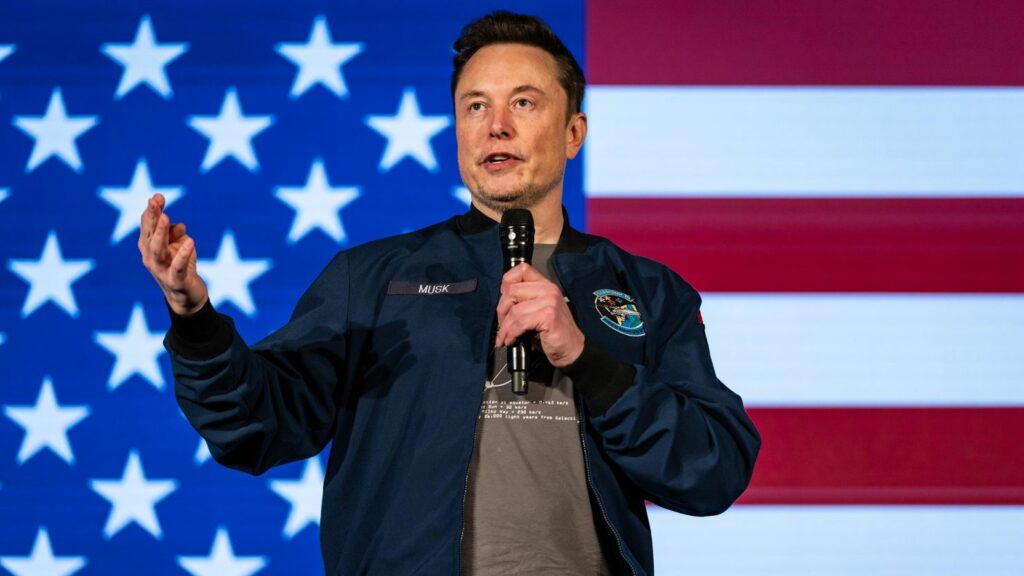 Elon musk holding a microphone and speaking in front of an american flag backdrop