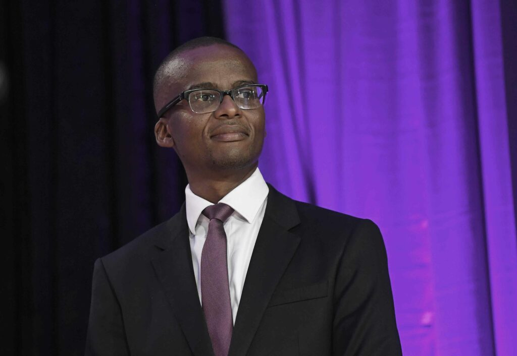 Ndaba gaolathe wearing glasses a dark suit and a purple tie looking confidently into the distance against a background illuminated with purple lighting