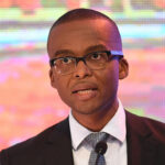 Ndaba gaolathe speaking at a podium wearing glasses and a suit with a patterned tie with a colorful background behind him