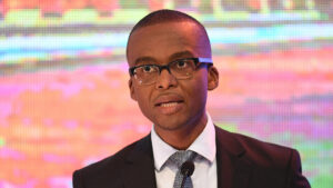 Ndaba gaolathe speaking at a podium wearing glasses and a suit with a patterned tie with a colorful background behind him