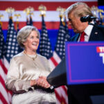 President Donald Trump shakes hands with Susie Wiles on stage during a 2024 campaign event, with American flags in the background.