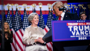 President donald trump shakes hands with susie wiles on stage during a 2024 campaign event with american flags in the background