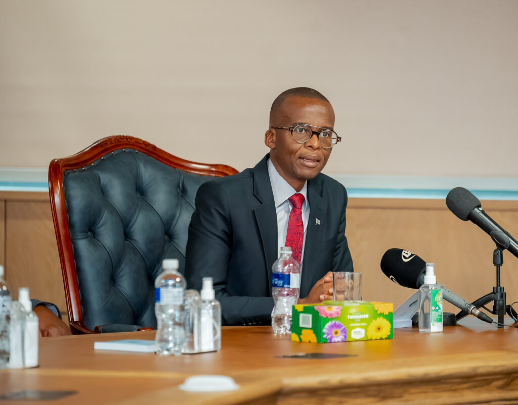 Vice president ndaba gaolathe of botswana speaking at the swearing in ceremony of new cabinet ministers seated at a table with a microphone and bottled water