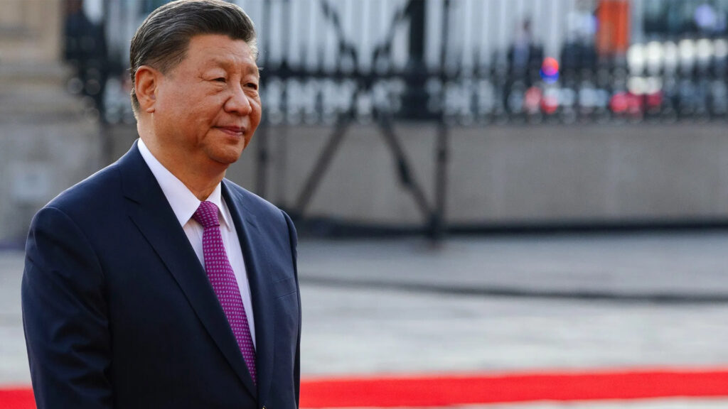 Chinese President Xi Jinping in a dark suit with a pink tie, walking along a red carpet at an official event.