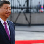 Chinese President Xi Jinping in a dark suit with a pink tie, walking along a red carpet at an official event.