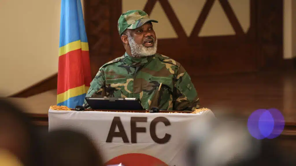 Corneille nangaa wearing a camouflage uniform speaks from behind a podium draped with the alliance fleuve congo afc insignia with the drc national flag in the background