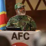 Corneille Nangaa, wearing a camouflage uniform, speaks from behind a podium draped with the Alliance Fleuve Congo (AFC) insignia, with the DRC national flag in the background.