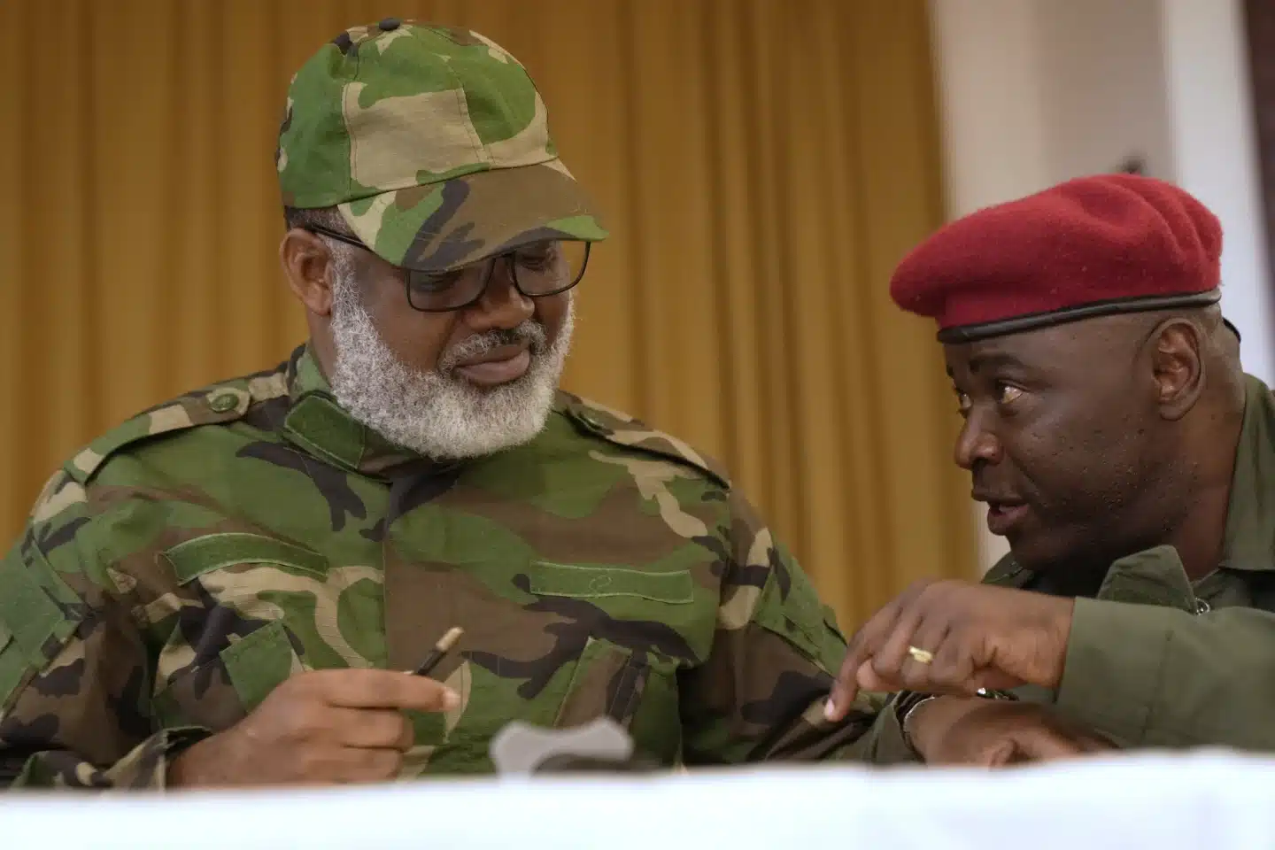 Corneille Nangaa, wearing camouflage military attire and glasses, consults with a red-beret military official at a meeting.