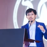 Liang Wenfeng, wearing a blue suit and glasses, speaks at a podium with two microphones, delivering a keynote speech on AI advancements.