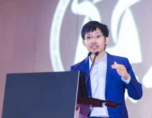 Liang wenfeng wearing a blue suit and glasses speaks at a podium with two microphones delivering a keynote speech on ai advancements