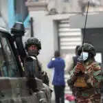 Armed M23 rebel fighters in camouflage uniforms and tactical gear stand near a military vehicle, one holding a radio for communication, amid ongoing clashes in Goma, DRC.