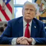 President Donald Trump in the Oval Office, dressed in a navy suit and red tie, making a statement on trade tariffs against Mexico, Canada, and China.