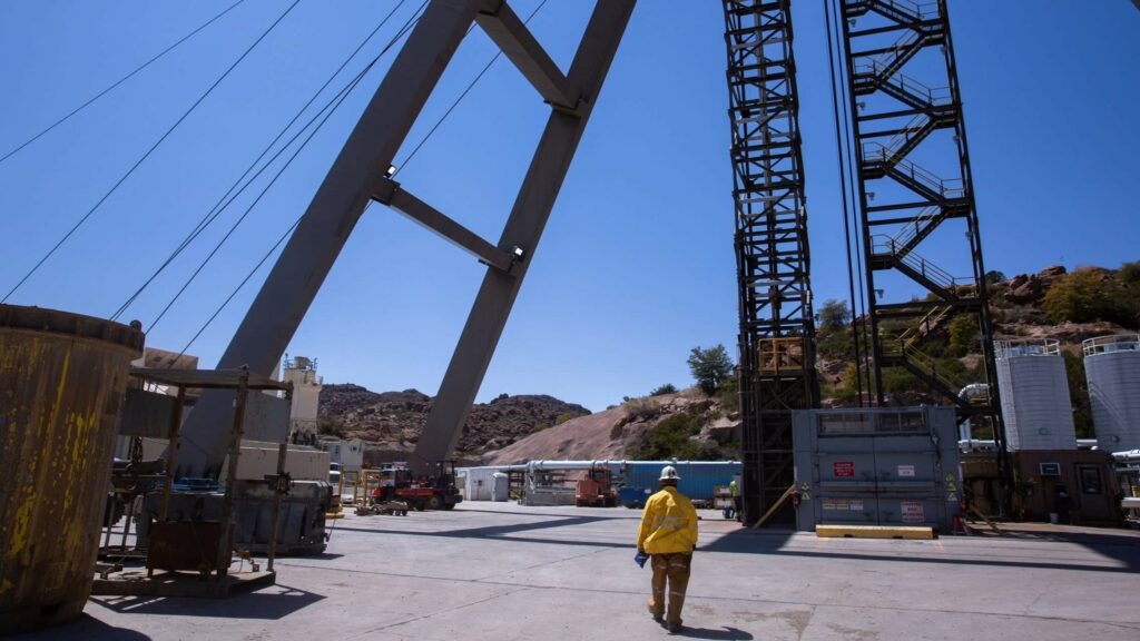 A worker in a yellow safety jacket walks through a copper mining site with large industrial structures cables and equipment against a desert like landscape