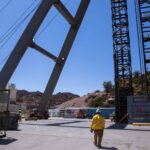 A worker in a yellow safety jacket walks through a copper mining site with large industrial structures, cables, and equipment against a desert-like landscape.