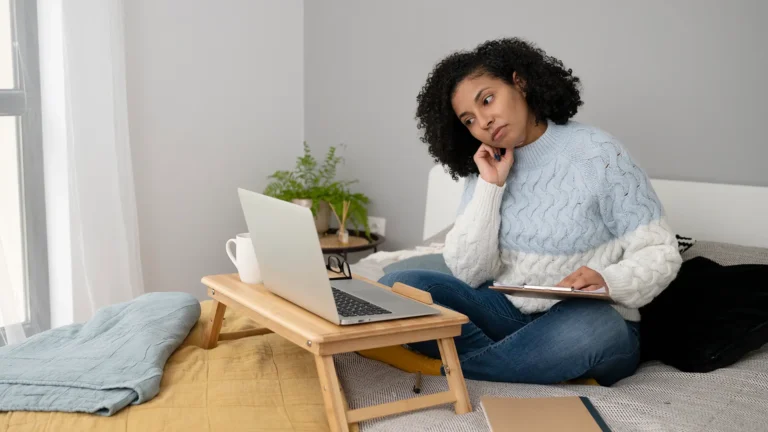 Woman in casual clothing sitting on a bed and attending a remote training session via laptop while taking notes.